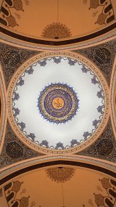 Ornate interior view of the central dome of Çamlıca Mosque in Istanbul, featuring intricate Islamic calligraphy and geometric patterns in gold, blue, and white, with the name of Allah in the center.