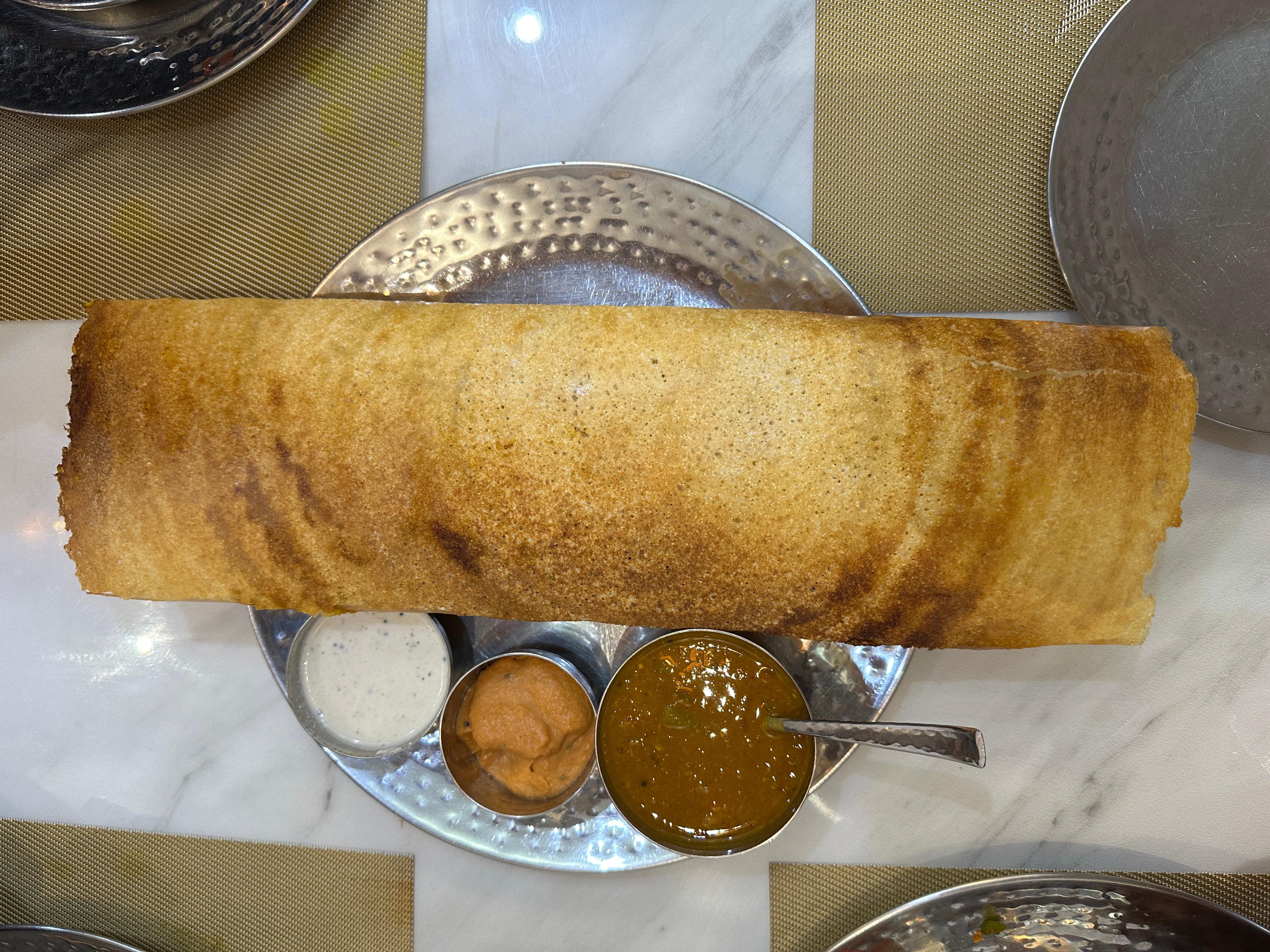 A very long, crispy dosa is served on a metal tray with three small bowls of different sauces for dipping. 