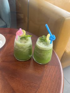 Two glasses of green mint lemonade, topped with lemon and mint—one with a pink straw, the other blue—sitting on a wooden table.