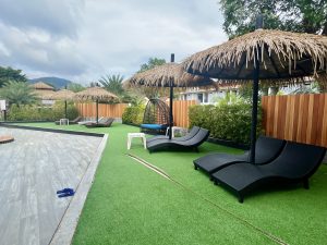 A serene outdoor space featuring a poolside area with artificial grass. There are two black lounge chairs positioned under thatched umbrellas, alongside a hanging chair with a blue cushion. Lush greenery and wooden fencing provide privacy in the background, with a cloudy sky above.