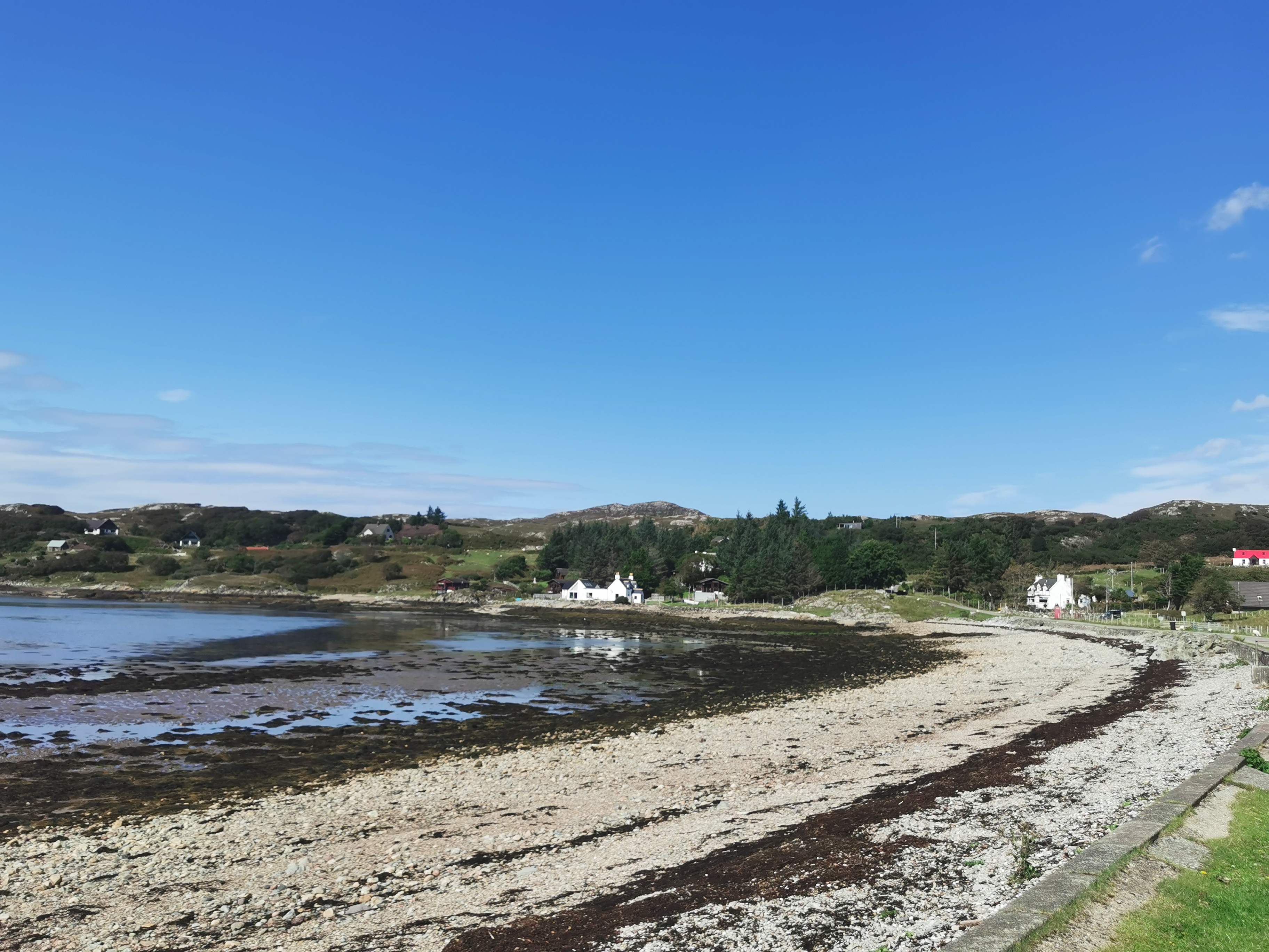 Rock and pebble shoreline, flanked by the sea to the left in the distance a white cottage sits at the base of a hill