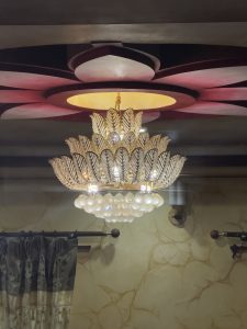 A decorative chandelier hanging from a ceiling featuring a floral design. The chandelier is composed of golden elements and numerous crystal-like accents that reflect light. The walls behind it are adorned with a textured finish in a light color, contributing to an elegant atmosphere.