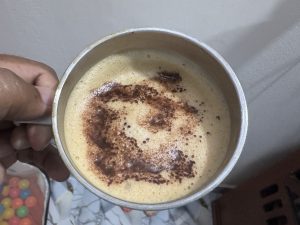 A person's hand holding a metal mug filled with frothy coffee, showing a swirl of dark coffee grounds on the surface.