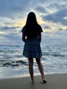 At dawn on a quiet shore, a person stands barefoot on wet sand, facing the sea. Their dark hair and patterned dress form a soft silhouette against a pale, cloud-streaked sky. Gentle waves curl in, reflecting the first light.