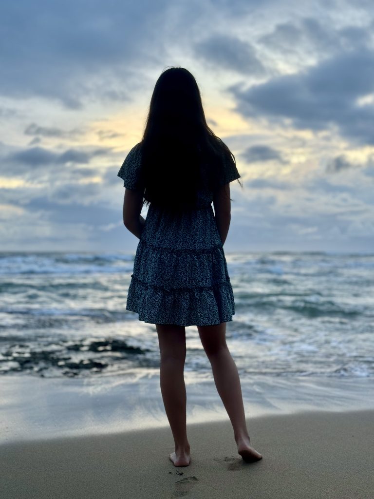 At dawn on a quiet shore, a person stands barefoot on wet sand, facing the sea. Their dark hair and patterned dress form a soft silhouette against a pale, cloud-streaked sky. Gentle waves curl in, reflecting the first light.