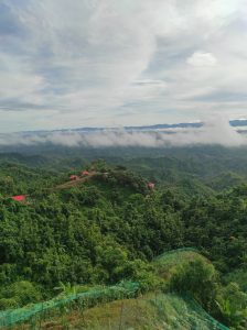 A panoramic view of a lush, green mountainous landscape under a cloudy sky. In the distance, a layer of clouds hovers above the lower valleys, while several small buildings with red roofs are situated on a hilltop amidst the greenery