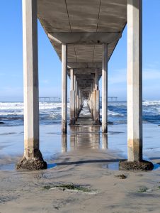 Underneath the Ocean Beach Pier in San Diego, CA. A forced-perspective view underneath concrete pier pylons with a wet sandy beach in the foreground and churning ocean waves in the background.