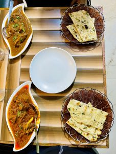A wooden table is set with a variety of dishes. On the left, a white, oval dish contains a rich, brown curry with pieces of meat, garnished with green herbs.
