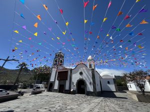A white church with a bell tower stands in a sunny plaza in Santiago del Teide (Tenerife-Spain), decorated with colorful triangular flags strung overhead against a deep blue sky, with mountains and palm trees in the background.