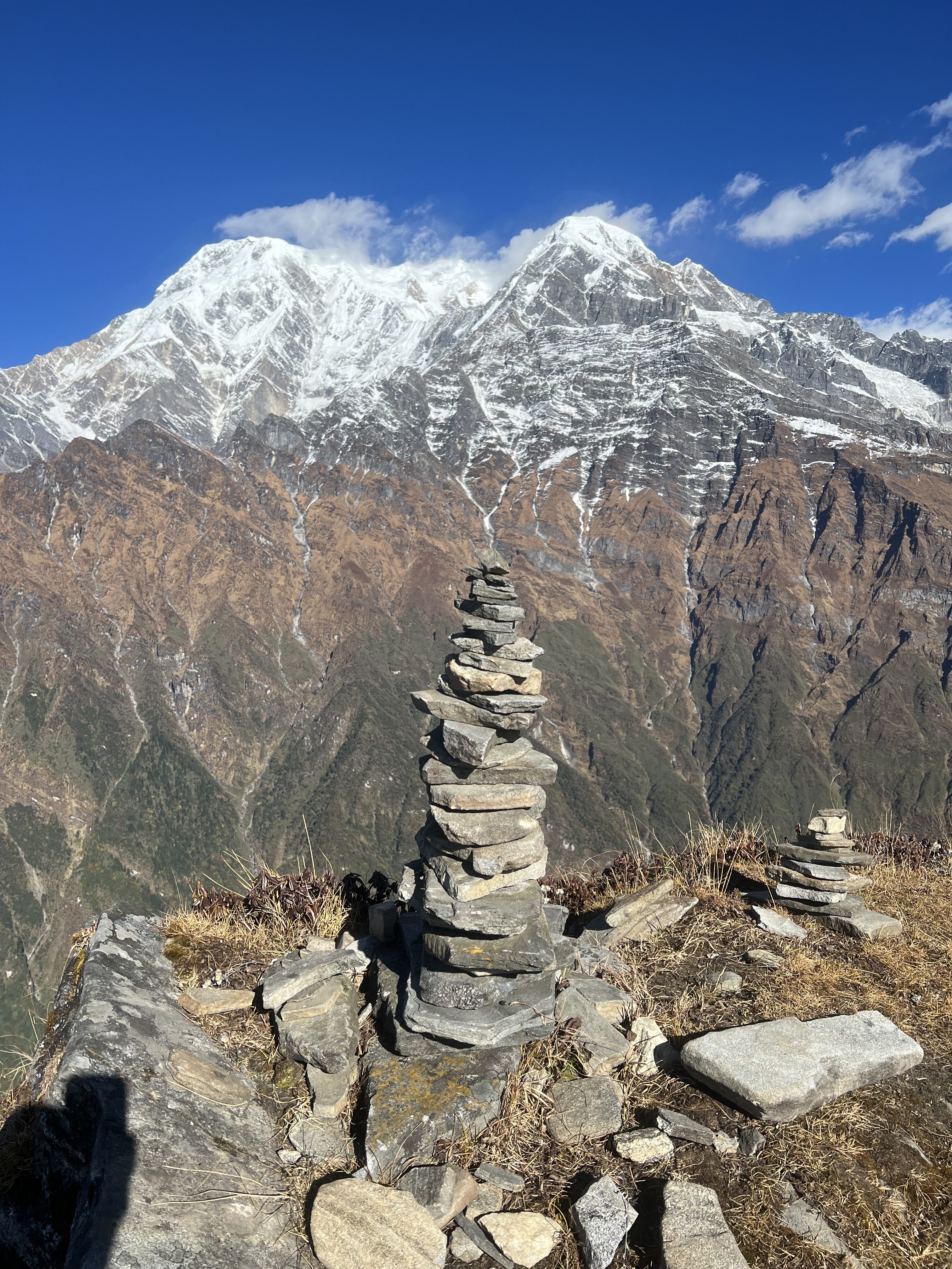 A tall, neatly stacked stone cairn stands on a rocky mountain ridge with a massive snow-capped mountain behind it.