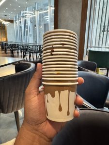 
A hand holds a neat stack of small paper cups with a chocolate-drip design, lightly smudged with real chocolate, against the backdrop of a softly lit modern restaurant.