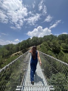 A girl wearing blue jumpsuit with long brown hair, seen from behind, walking across a metal suspension bridge over a forested valley on a sunny day.