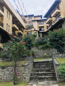 Vibrant prayer flags hang above a courtyard with tiered landscaping, stone walls, and stairs leading up to a resort-style building complex. 