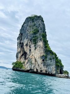 A very tall, steep rock formation covered in green foliage rising out of the turquoise sea under a cloudy sky.