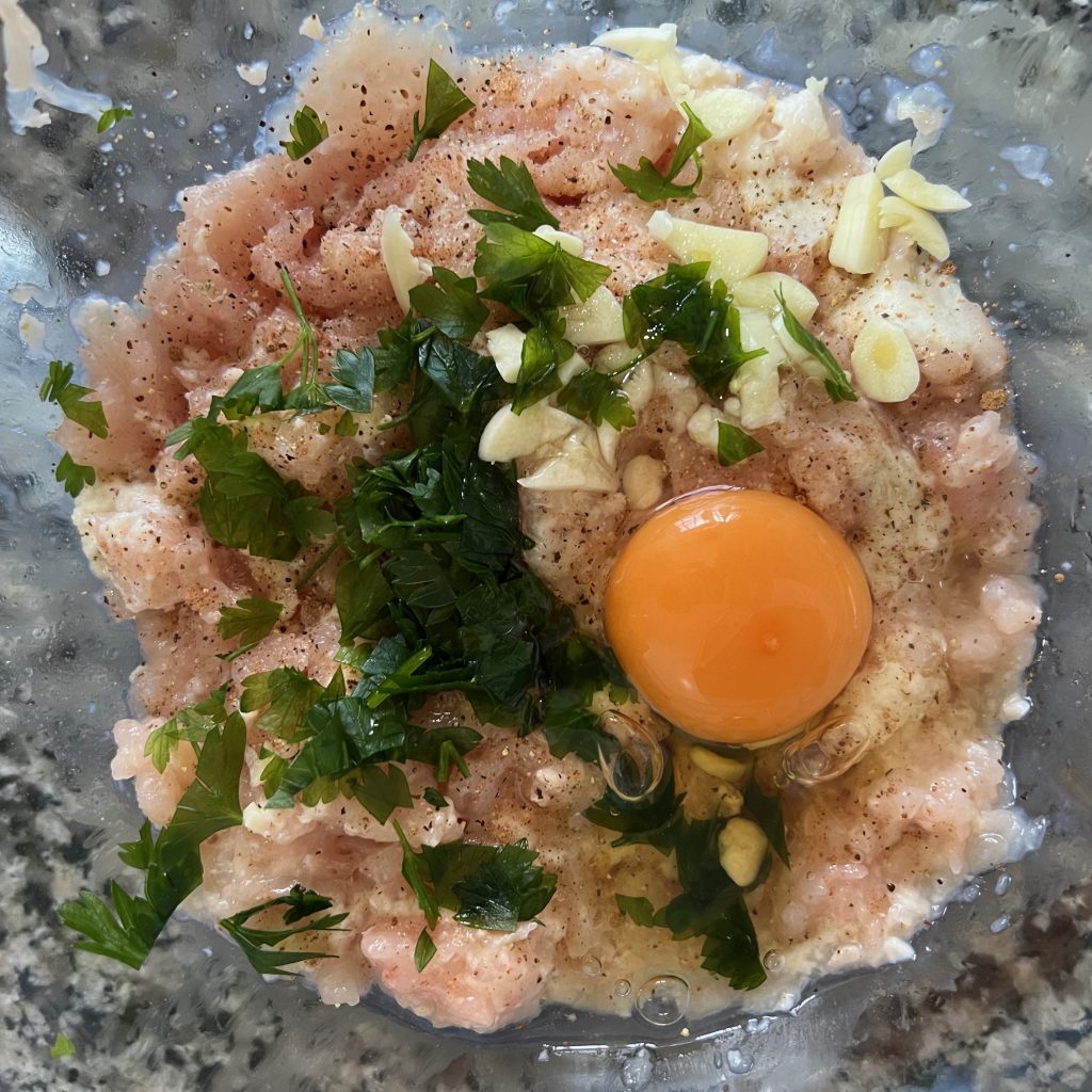 A glass bowl of minced chicken topped with a raw egg, parsley, garlic, and black pepper on a dark countertop