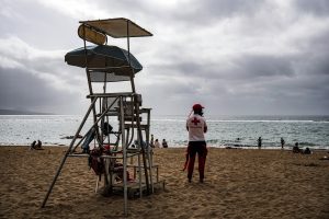 A lifeguard on duty stands on a sandy beach, overlooking the ocean. The lifeguard wears a red hat and a shirt with a red cross, symbolizing their role in safety. In the background, a lifeguard tower is visible, along with beachgoers enjoying the water and sand under a cloudy sky. 