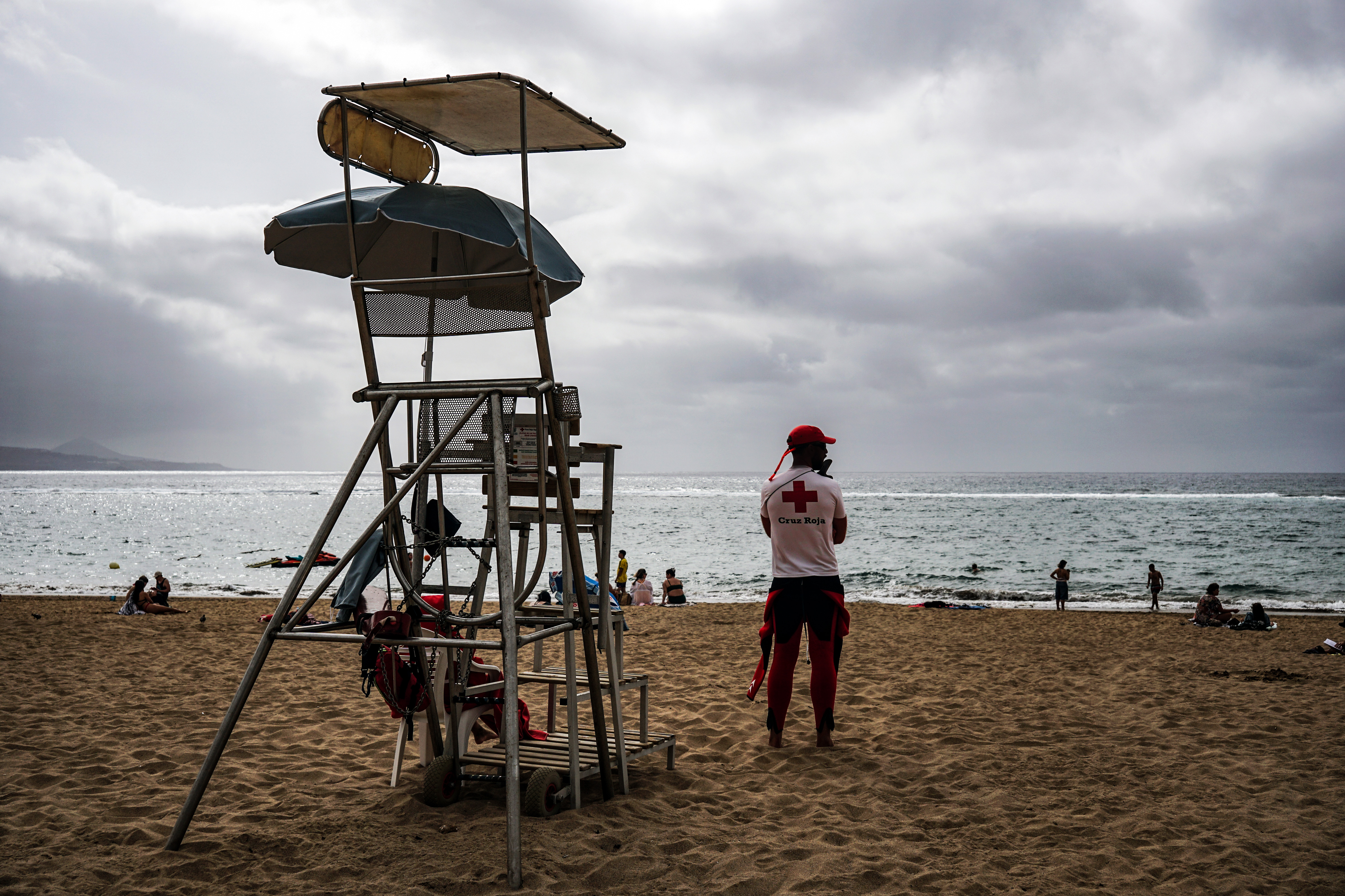 A lifeguard on duty stands on a sandy beach, overlooking the ocean. The lifeguard wears a red hat and a shirt with a red cross, symbolizing their role in safety. In the background, a lifeguard tower is visible, along with beachgoers enjoying the water and sand under a cloudy sky.