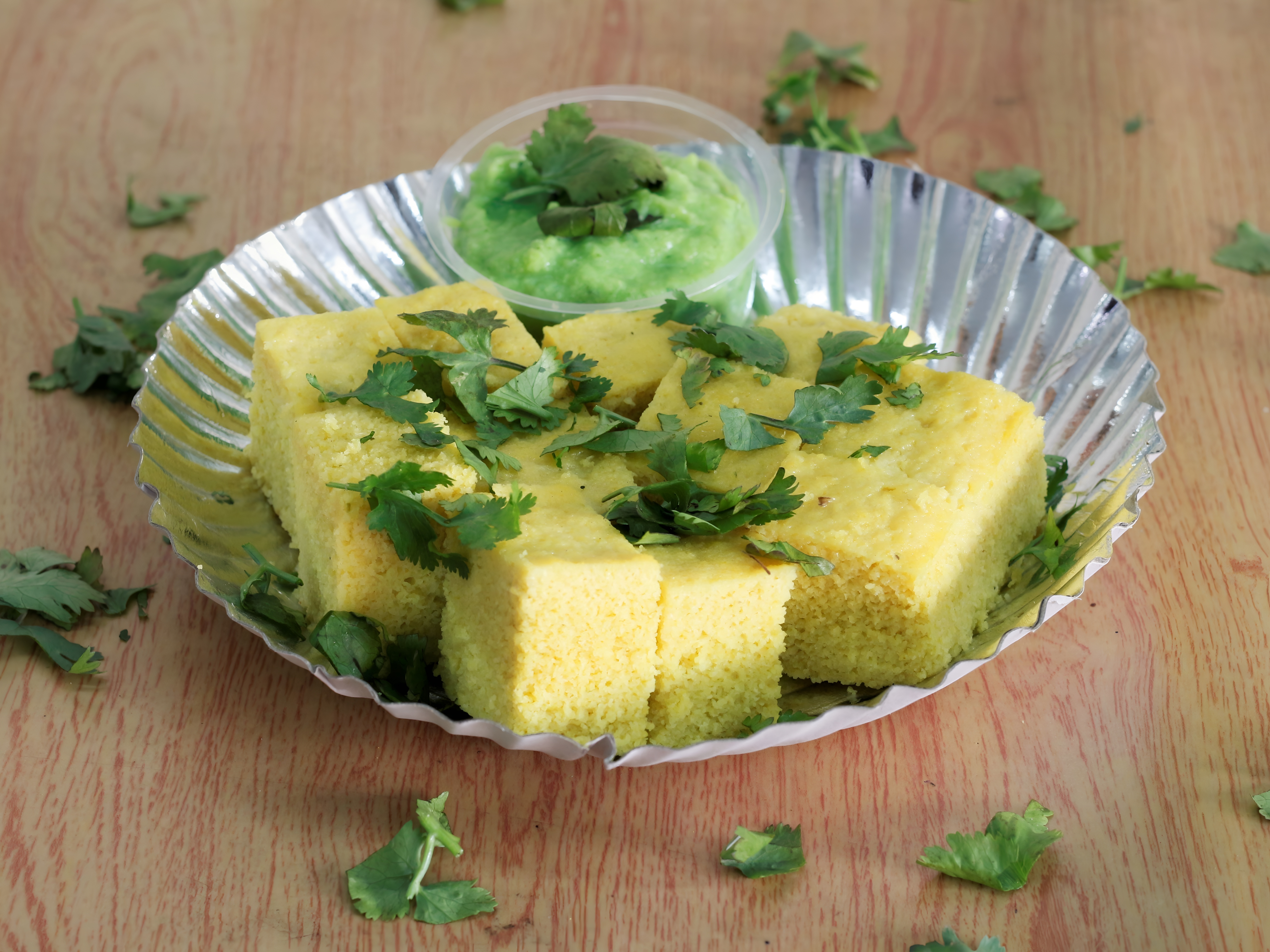 A serving of soft yellow Khaman Dhokla garnished with fresh coriander leaves, placed in a silver foil plate with a small container of green chutney, on a wooden table.
