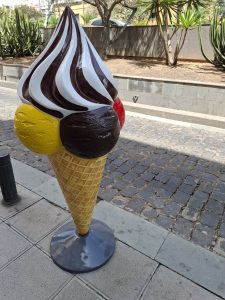 Ice cream sculpture with a cone topped by yellow, brown, and red scoops, crowned with a white-and-brown whipped cream swirl. 