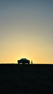 A silhouette of a man walking towards a truck on a sandy landscape at sunset. The sky is a gradient of blue to soft yellow, blending into the horizon.