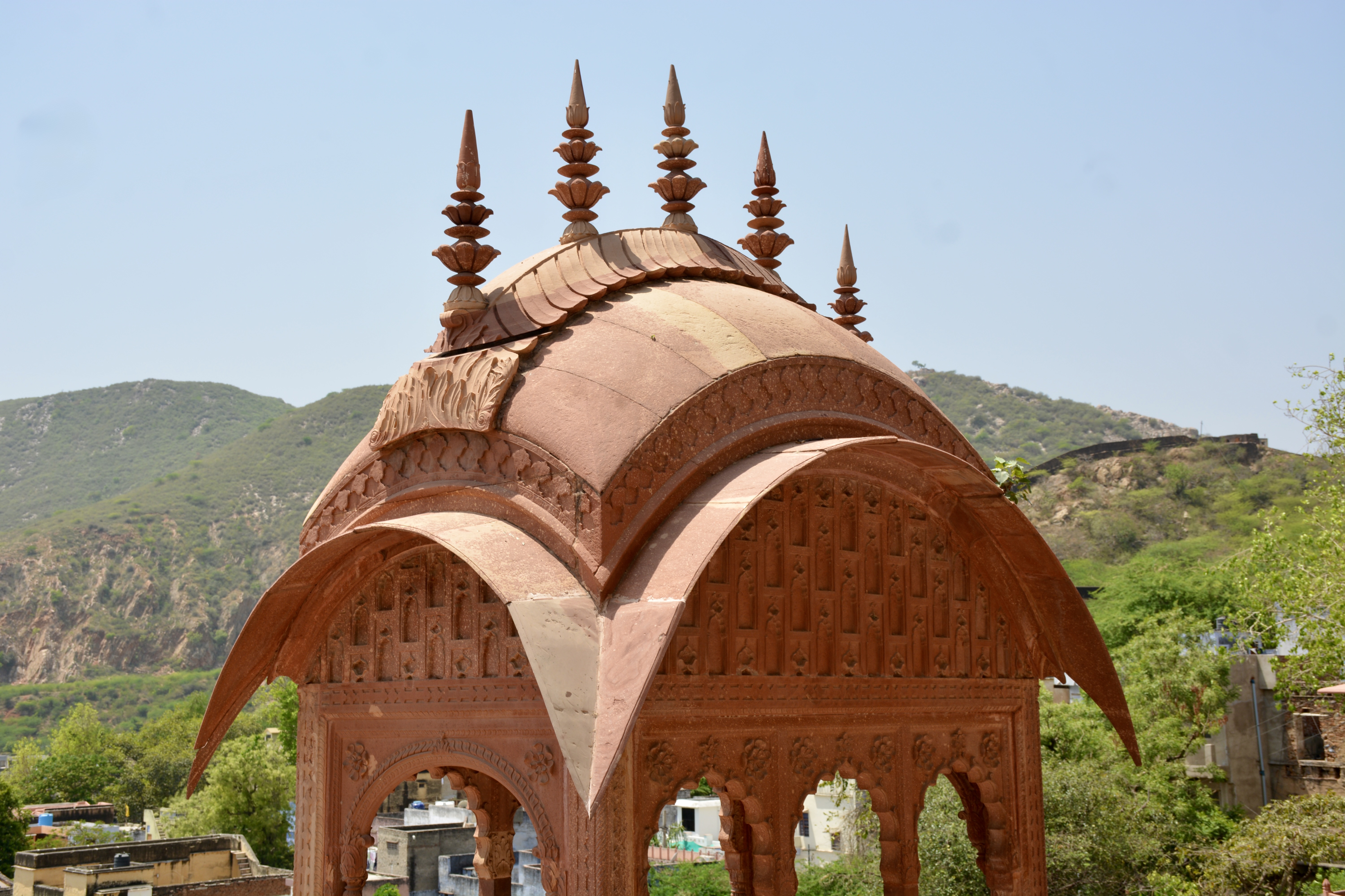 Close-up of an ornate red sandstone chhatri roof at Moosi Maharani Ki Chhatri, Alwar, Rajasthan, adorned with floral finials and set against the backdrop of green hills and the Bala Quila fort wall.