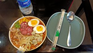 ramen topped with boiled eggs, sausage slices, sweet corn, green onions, mushrooms, meat slices, and thick noodles in a rich-looking broth, garnished with sesame seeds. Beside it, there’s a green plate with a spoon and a pair of chopsticks in a wrapper