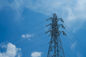 A tall steel electricity transmission tower with multiple power lines stretching across a bright blue sky with scattered white clouds.
