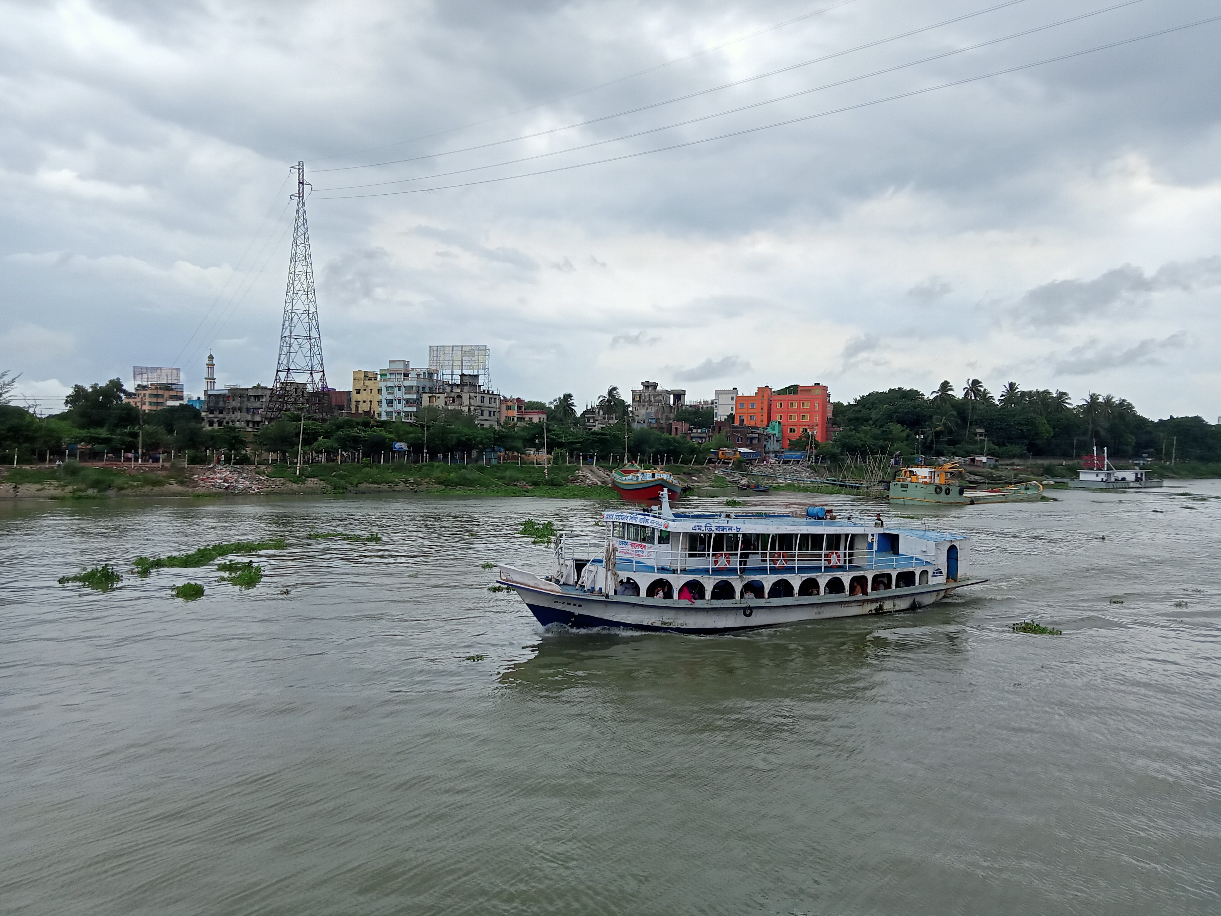 A river scene featuring a white and blue passenger boat navigating through the water. In the background, a variety of buildings and structures line the riverbank, including an antenna tower and colorful houses.