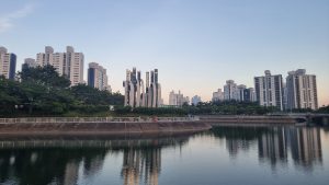 Daytime view of Daecheon Lake in Daecheon Park, Haeundae, Busan, surrounded by green trees and high-rise apartments, with modern sculptures standing prominently in the center.

