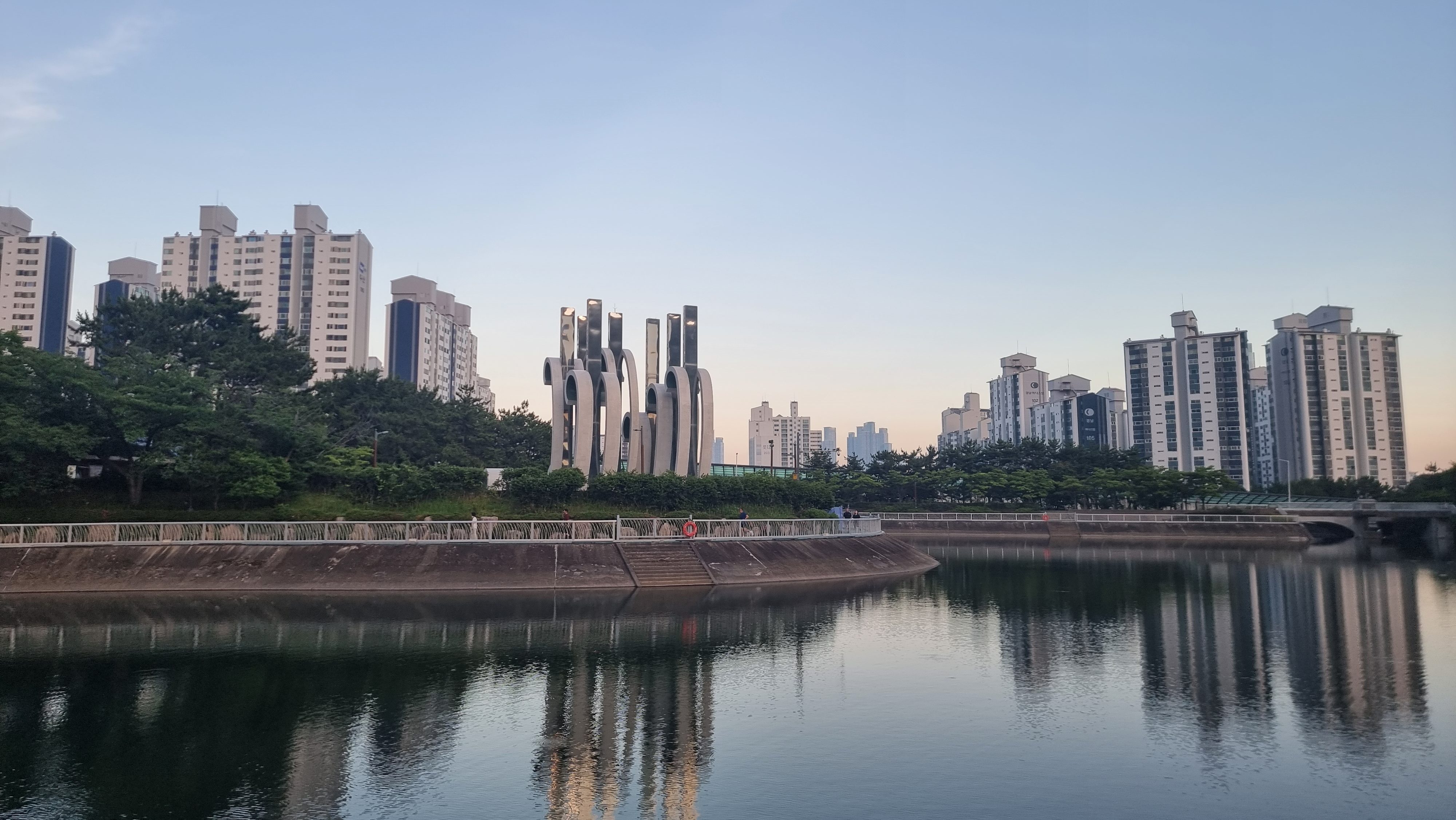 Daytime view of Daecheon Lake in Daecheon Park, Haeundae, Busan, surrounded by green trees and high-rise apartments, with modern sculptures standing prominently in the center.