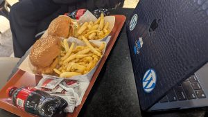 Two burgers with fries and a drink behind a laptop with dark blue patterned cover with WordPress Mark stickers on the cover bottom.