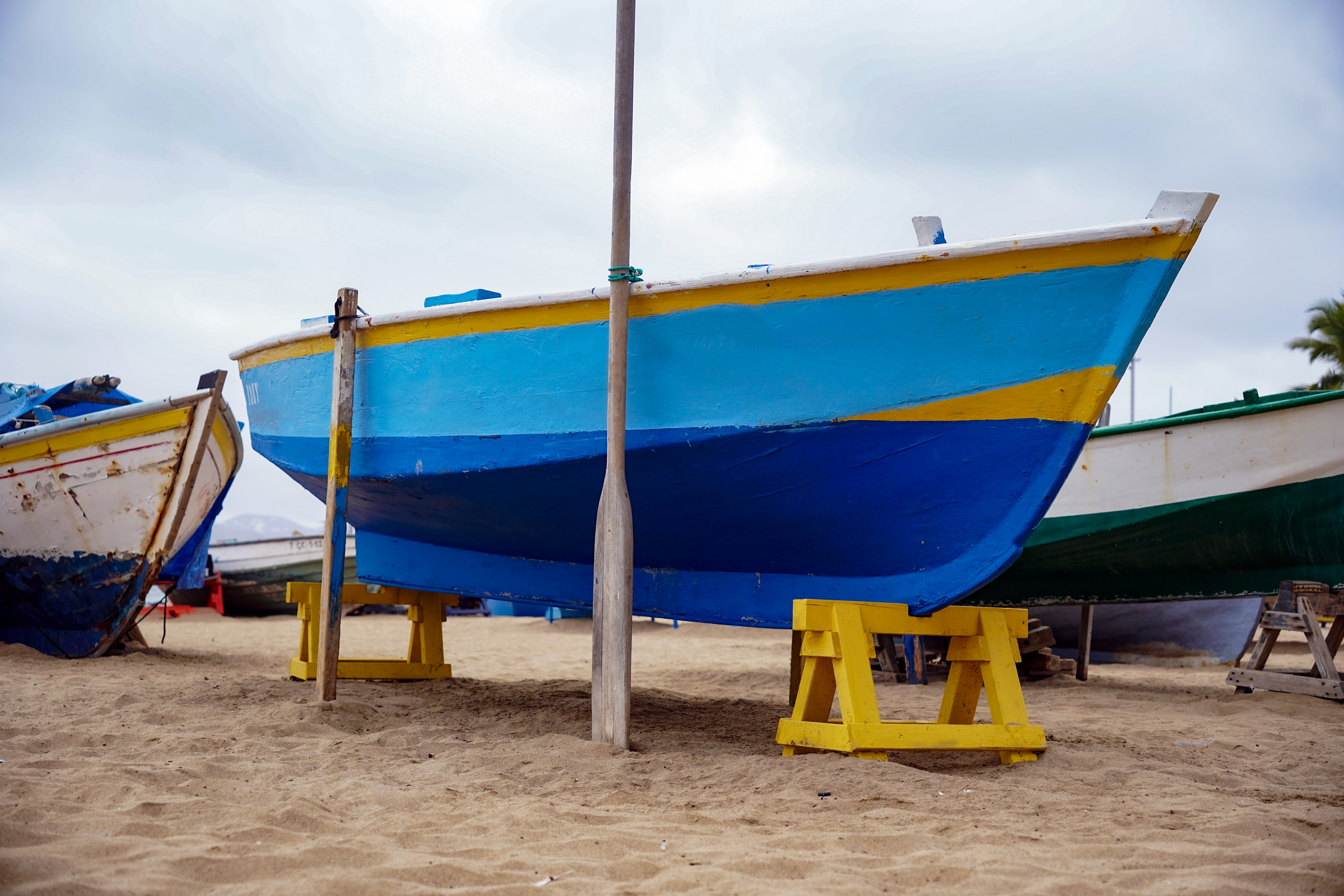 A brightly painted blue fishing boat with yellow accents is propped up on yellow wooden supports on a sandy beach.