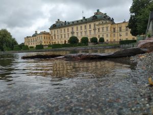 A low-angle shot from the water’s edge capturing Drottningholm Palace in Sweden under a cloudy sky. The palace, a large, light-colored building with green roofs, stands prominently in the background, reflected slightly in the calm water of the lake or canal in the foreground. Rocks and small pebbles are visible along the shoreline in the immediate 
