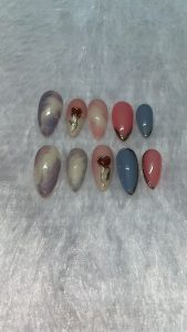 A collection of ten artificial nails displayed on a soft, light gray fabric surface. The nails feature various designs and colors: three are marbled in pastel hues of blue, pink, and beige, accented with a golden heart design. The others are solid colors in pink, light blue, and two shades of peach, with some having a glossy finish and gold detailing along the edges.
