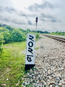 A white railway marker post with Bengali script next to the track, with a red signal light in the distance.
