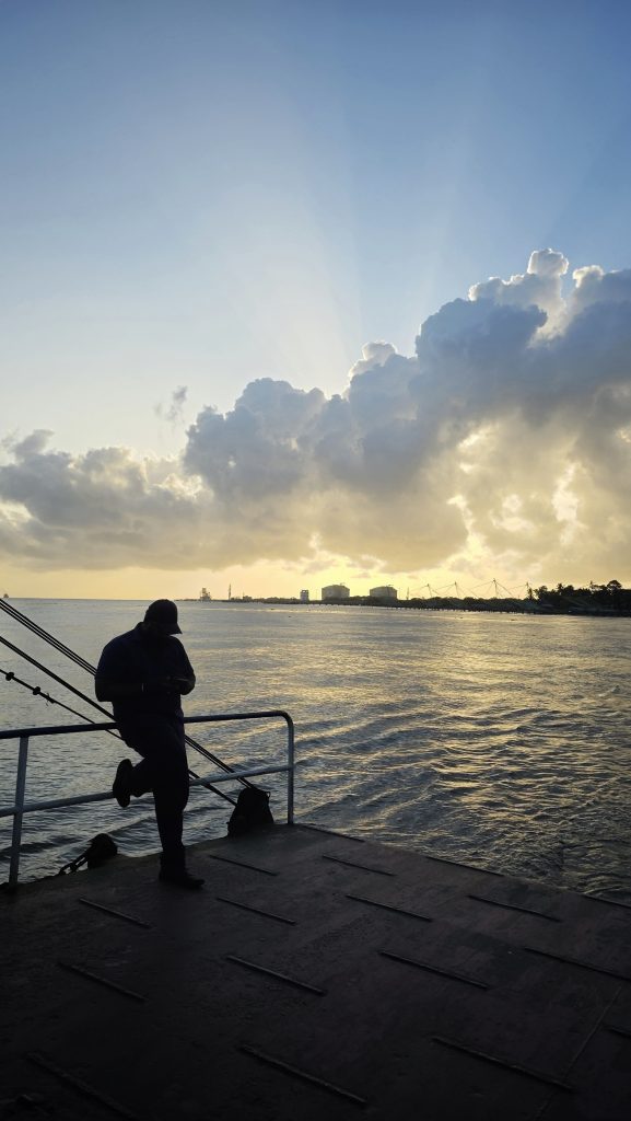 A silhouetted man stands on the deck of a Ro-Ro, looking out at a calm body of water with a city skyline in the distance under a dramatic sunset sky with clouds having the chinese fishing nets. Photo taken from Kochi, Kerala.