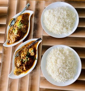 A wooden table with two oval dishes of spicy meat curry garnished with green herbs and two plates of fluffy white rice.