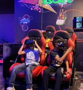 Children immersed in a thrilling VR adventure, surrounded by vibrant lights and futuristic chairs, enjoying a summer gaming escape