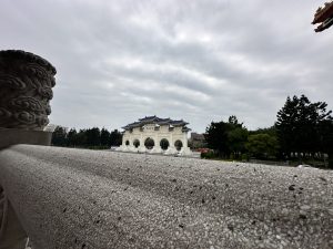 The Gate of Great Loyalty at Chiang Kai-shek Memorial Hall from the side, with a stone railing in the foreground and a cloudy sky above the white gate with its blue roof.