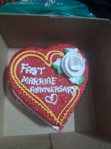 Heart-shaped red cake inside a brown paper box with "Fast Marriage Anniversary" written in white icing.