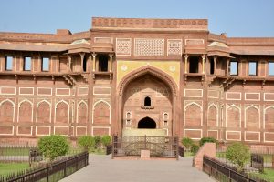 A close-up view of a historic red sandstone structure featuring intricate architectural details. The building has large arched doorways and windows adorned with ornate carvings. A decorative railing surrounds a stone basin situated in front of the entrance, with well-maintained greenery and pathways leading up to the structure against a clear blue sky.