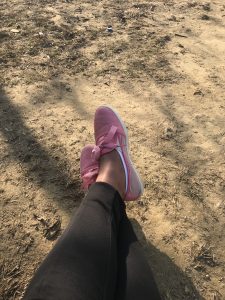 A person's legs are shown from the knees down, with pink sneakers on, resting on a dirt path with some dry leaves.