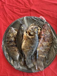 A round plate covered with plastic wrap holds four grilled fish. The fish are arranged closely together, with their heads facing the same direction. The background features a vibrant red tablecloth, which contrasts with the darker tones of the fish.