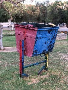 A worn, colorful, red-and-blue metal trash container sits on a grassy area, supported by a rusty, multicolored metal structure. Trees and park benches are partially blurred in the background.
