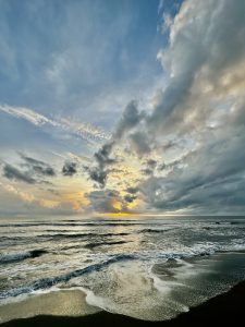 Dawn over a wild shore, where waves trace white foam on a dark beach and golden light breaks through heavy clouds into a blueening sky.