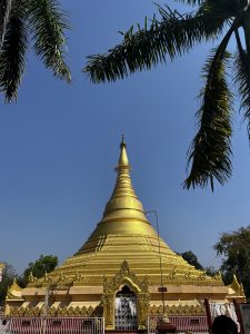 A golden stupa shines brightly under a clear blue sky, partially framed by the fronds of two palm trees. The stupa has a tiered design and an ornate golden spire at the top. 