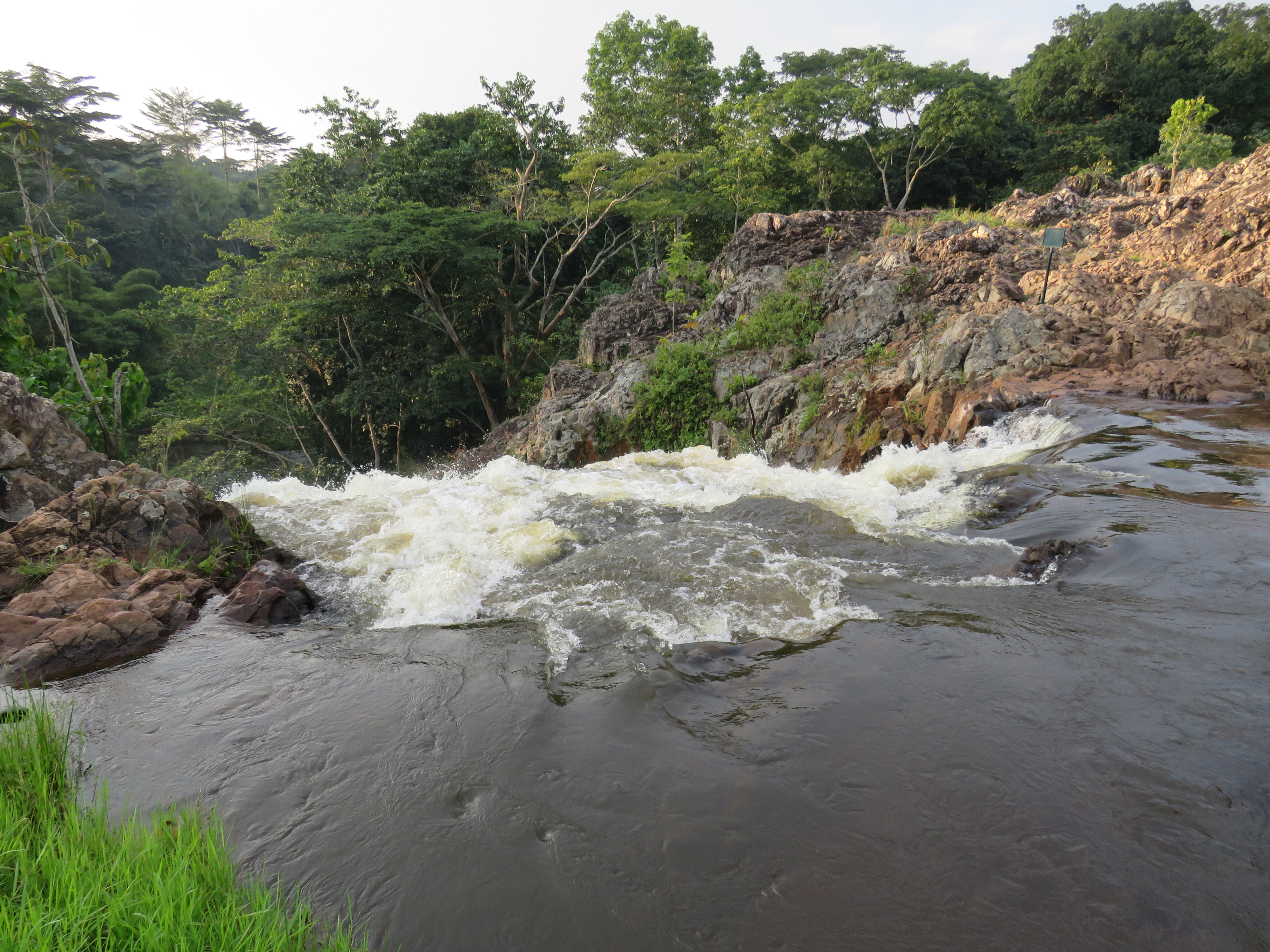 A rushing waterfall flows over rocky terrain, surrounded by lush green vegetation and trees.
