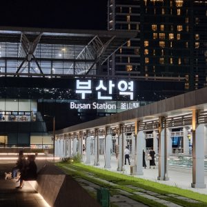 Night view of the illuminated Busan Station building, showing bright signage in Korean and English, with people walking and sitting outside in a modern plaza; tall apartment buildings rise in the background
