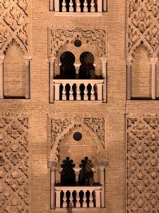 Historic building facade with stone carvings, decorative arches, and white-railed balconies separated by ornate wall designs. Textured brick backdrop showcases a blend of Moorish and Renaissance styles with geometric patterns and rich ornamentation.