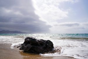 A rocky formation sits on the sandy beach, partially surrounded by gentle waves. The ocean stretches out towards the horizon, where light reflects off the water under a cloudy sky. 